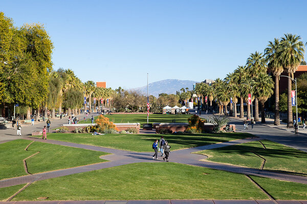 A view of the University of Arizona campus in Tucson features a mountain in the background, and students walking on paths framed by trees. 