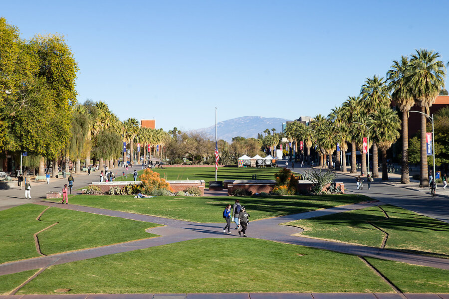 A view of the University of Arizona campus in Tucson features a mountain in the background, and students walking on paths framed by trees. 