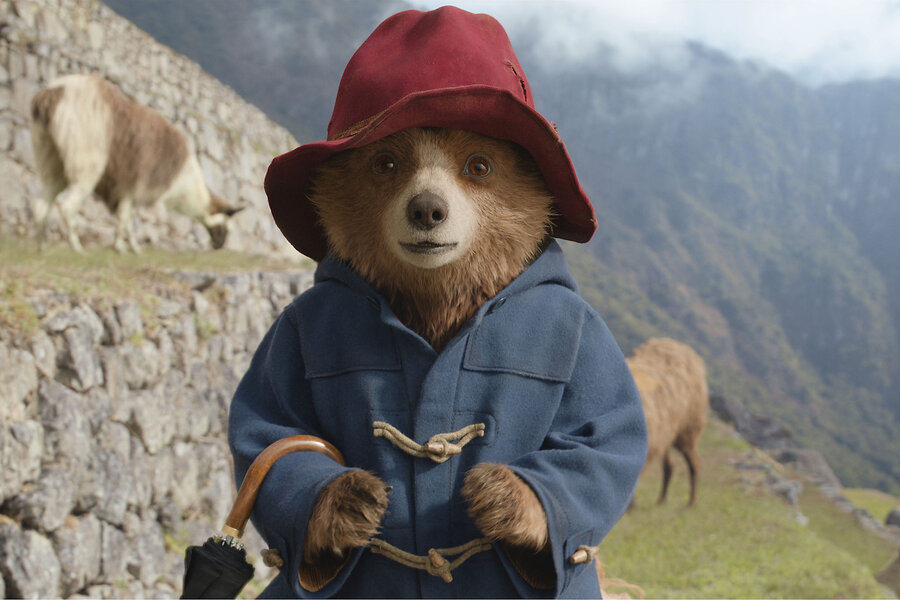 Paddington the bear stands looking at the camera wearing a red hat with mountains and animals behind him