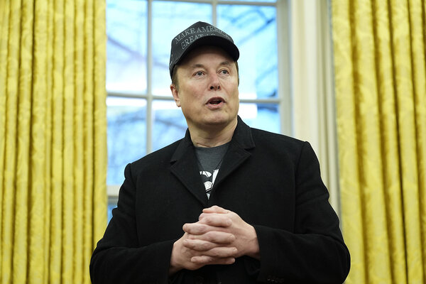 Elon Musk, wearing a baseball-style cap and dark jacket, speaks with an Oval Office window behind him.