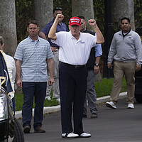 President Trump, wearing a red MAGA hat and white shirt, raises his fists above his head in a victory gesture.