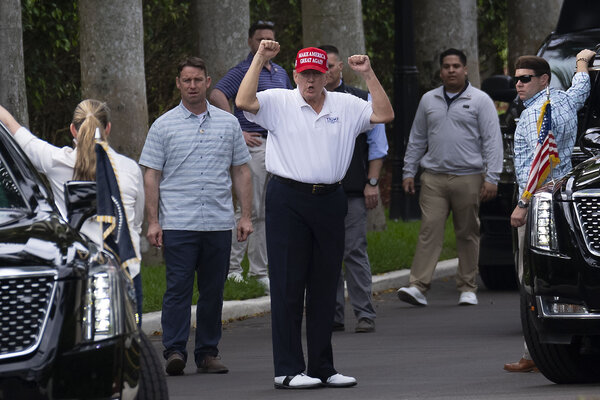 President Trump, wearing a red MAGA hat and white shirt, raises his fists above his head in a victory gesture.