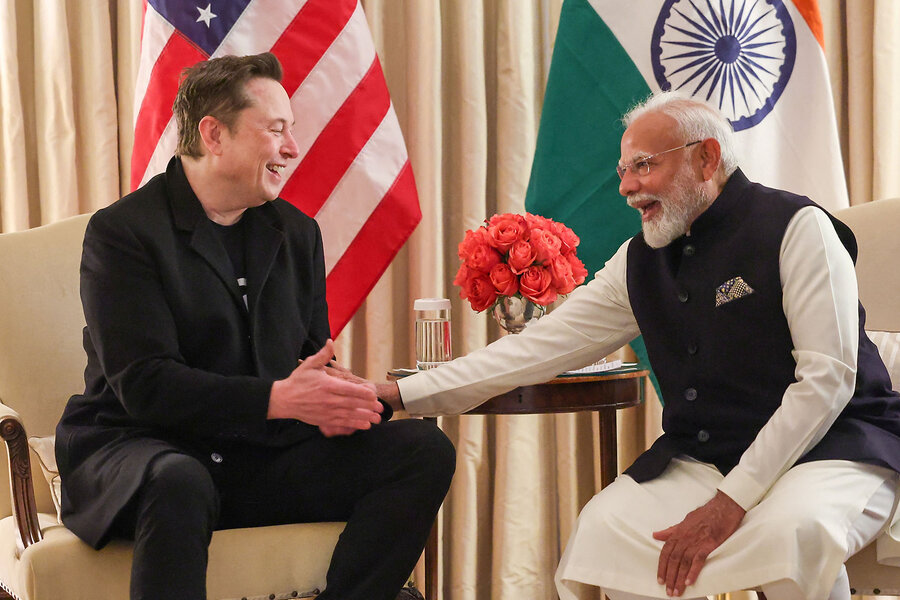 Elon Musk and Narendra Modi smile and shake hands, seated in a room with the flags of the United States and India behind them.