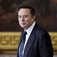 Elon Musk looks at the camera, wearing a dark suit and tie.