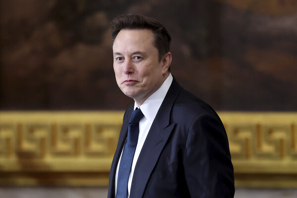 Elon Musk looks at the camera, wearing a dark suit and tie.