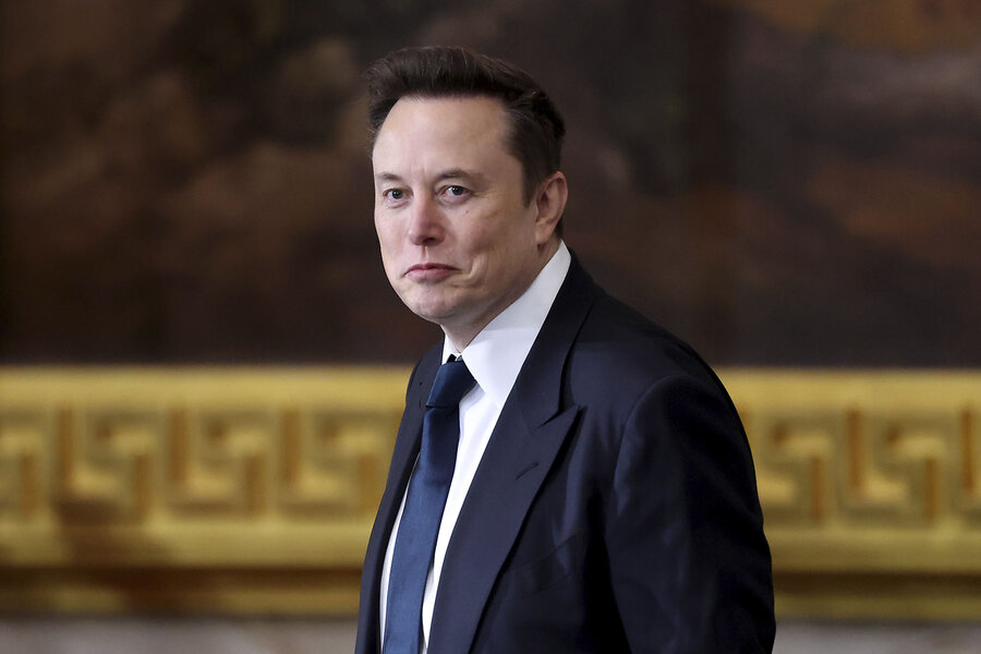 Elon Musk looks at the camera, wearing a dark suit and tie.