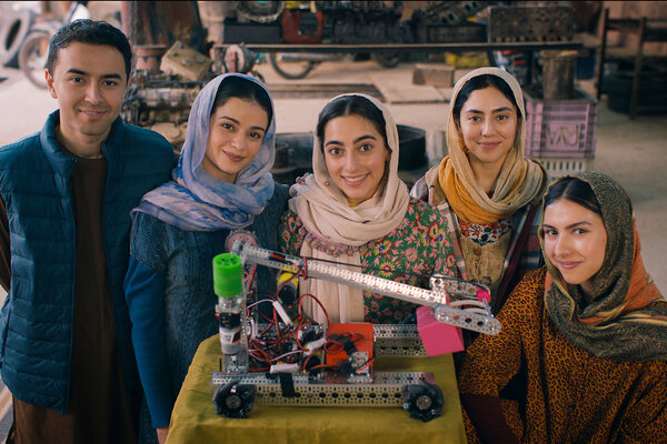 Actors in a film about the Afghan girls robotics team stand behind a robot on a table and face the camera smiling