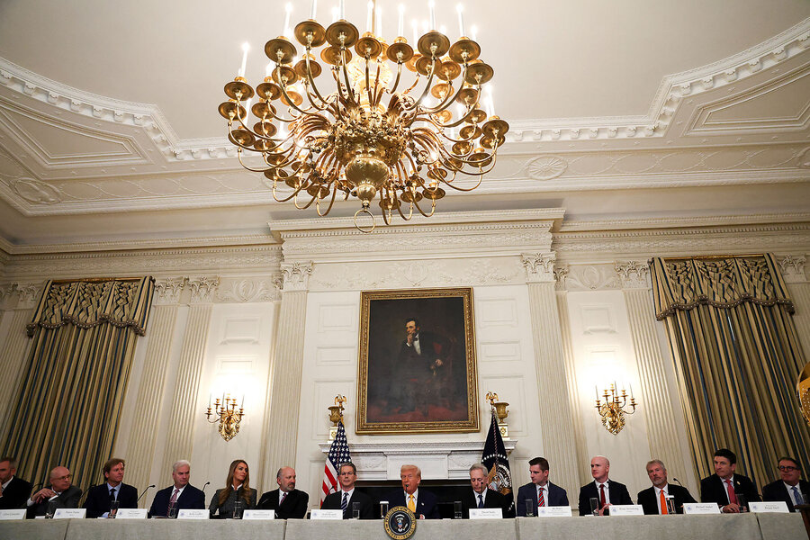 A dozen people in dark suits sit, with President Trump in the middle, one one side of a long table at the White House.