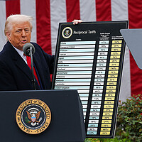 Donald Trump stands at a podium, showing a chart that lists tariffs for different nations. Behind him is large American flag.
