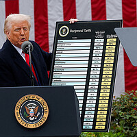 With a giant flag behind him, President Donald Trump stands beside a chart listing global tariffs in the Rose Garden.