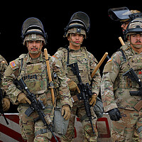 Members of the California National Guard wearing camoflage uniforms, riot helmets and rifles, stand together.