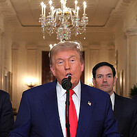 President Donald Trump with a red tie and blue suit speaks into a microphone in the White House.
